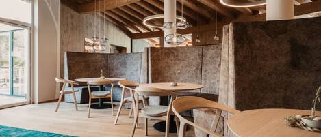 A modern dining area with wooden tables and chairs, a blue rug, and a high ceiling with exposed beams and pendant lighting.