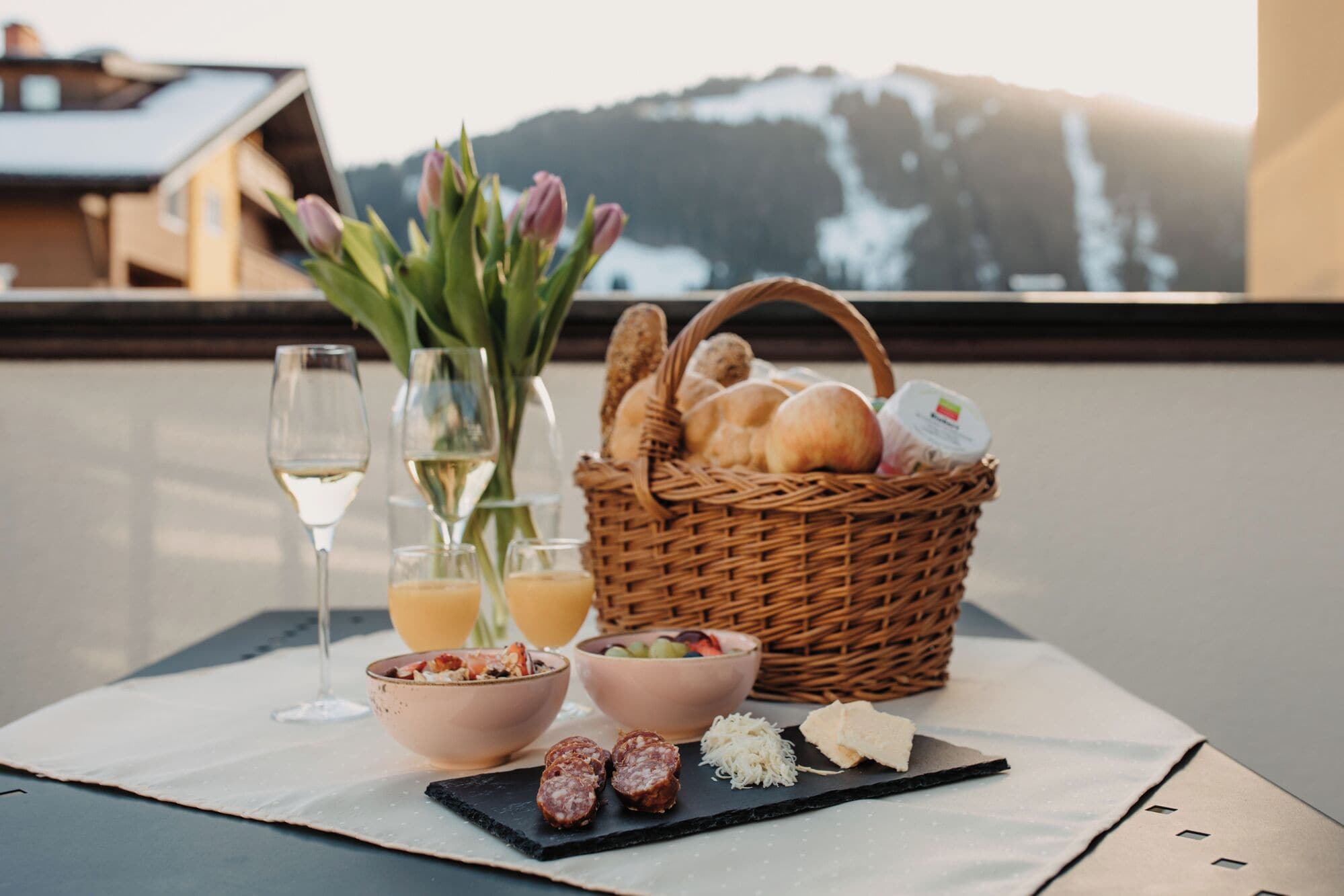 A table with a wicker basket containing bread, a bowl of fruit, and a glass of white wine.