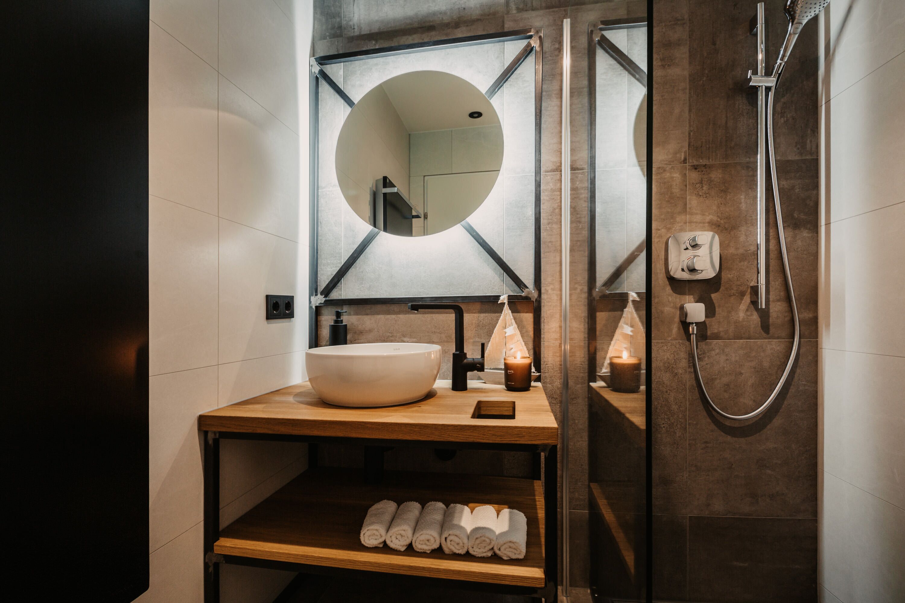 Een moderne badkamer met een grote ronde spiegel, een wastafel, een houten plank met handdoeken en een douche met een handdouche.
