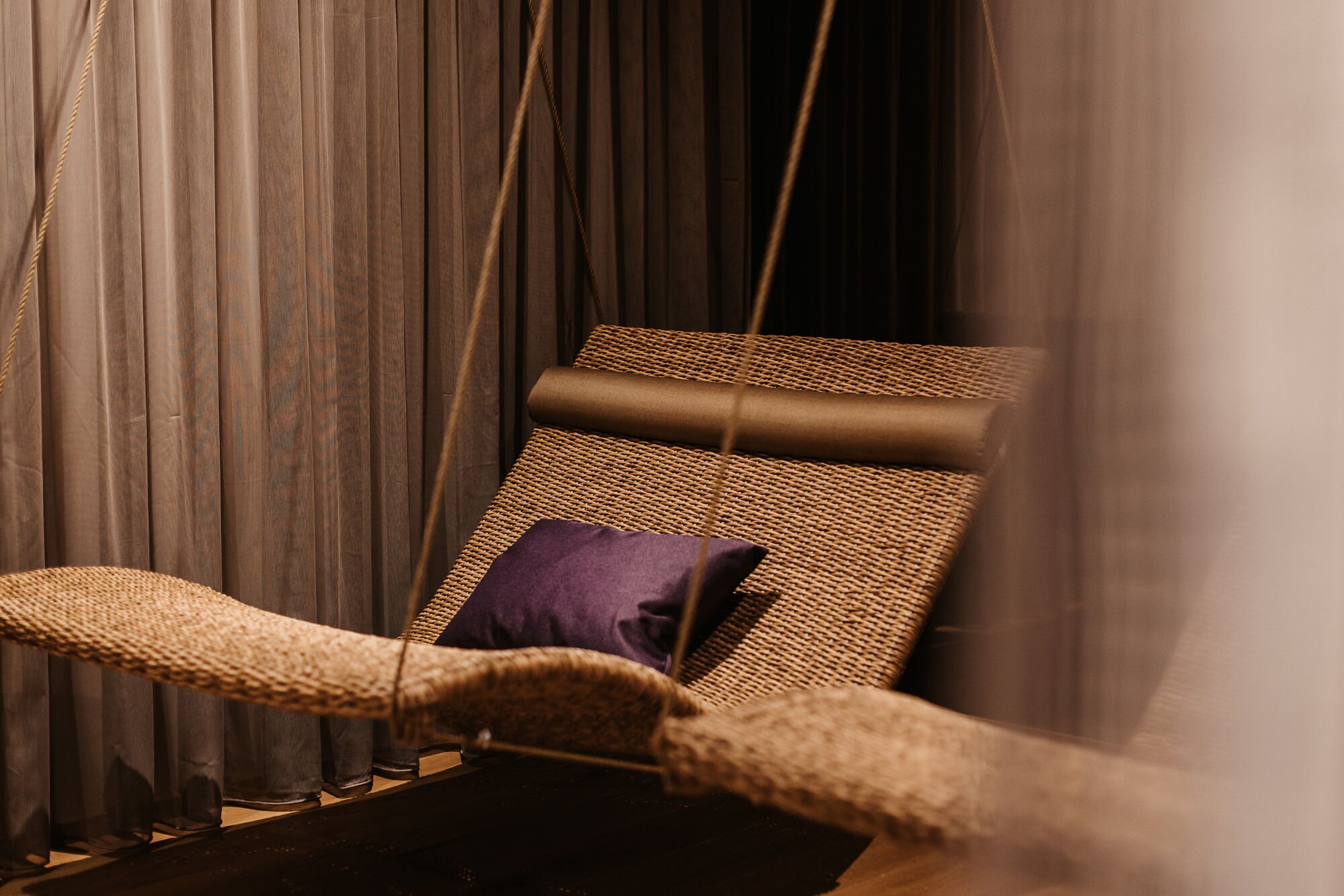 Una silla de mimbre con cojín morado, frente a cortinas oscuras.