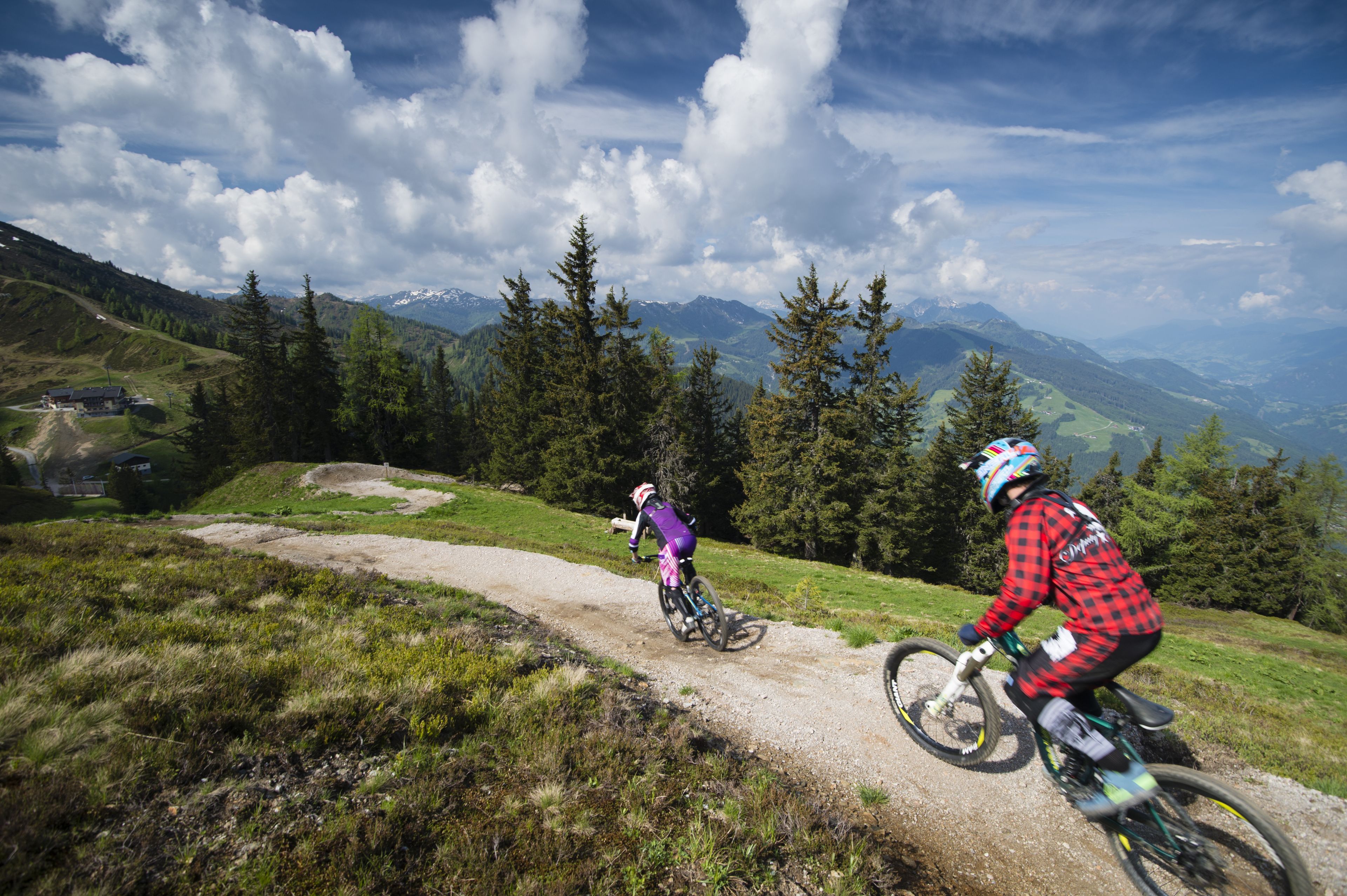Two mountain bikers on a dirt trail with a mountainous landscape in the background.