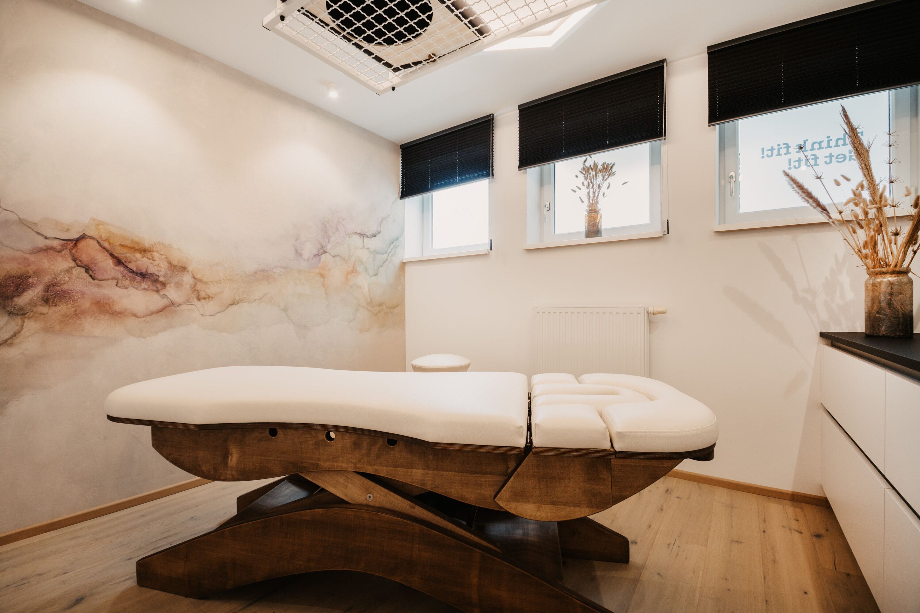 A modern treatment room with a wooden examination table, a marble wall, and a window with a view of the sky.