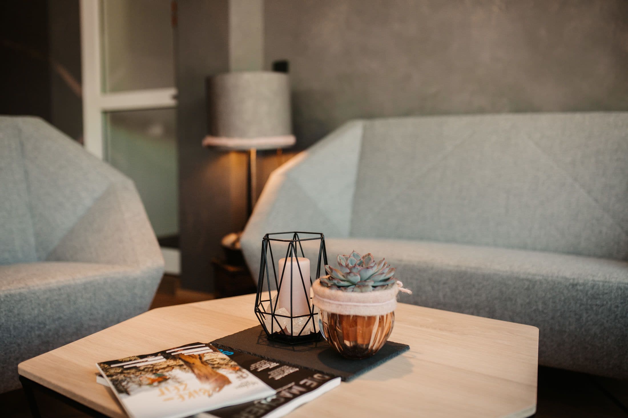 A modern living room with a light grey sofa, a wooden coffee table with a magazine, a candle in a geometric holder, and a succulent plant in a copper pot.