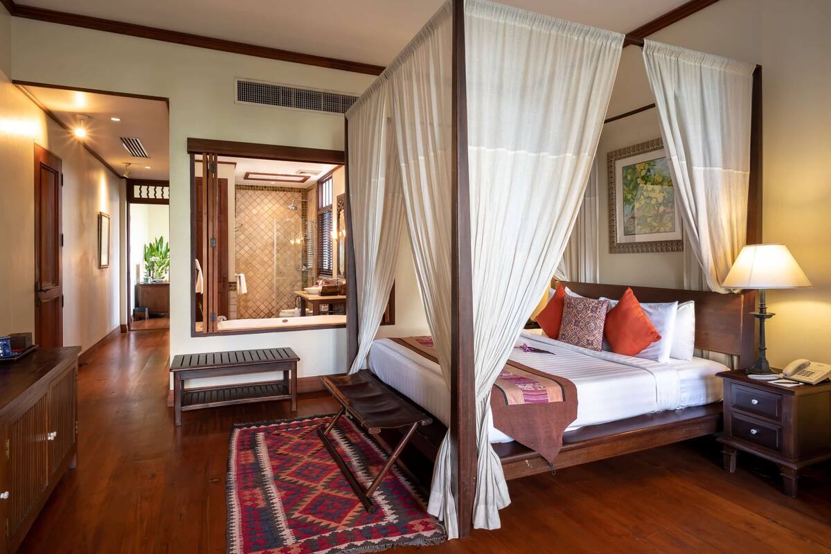 A four-poster canopy bed with a bench, a bedside table with a lamp, and a nightstand with a phone.