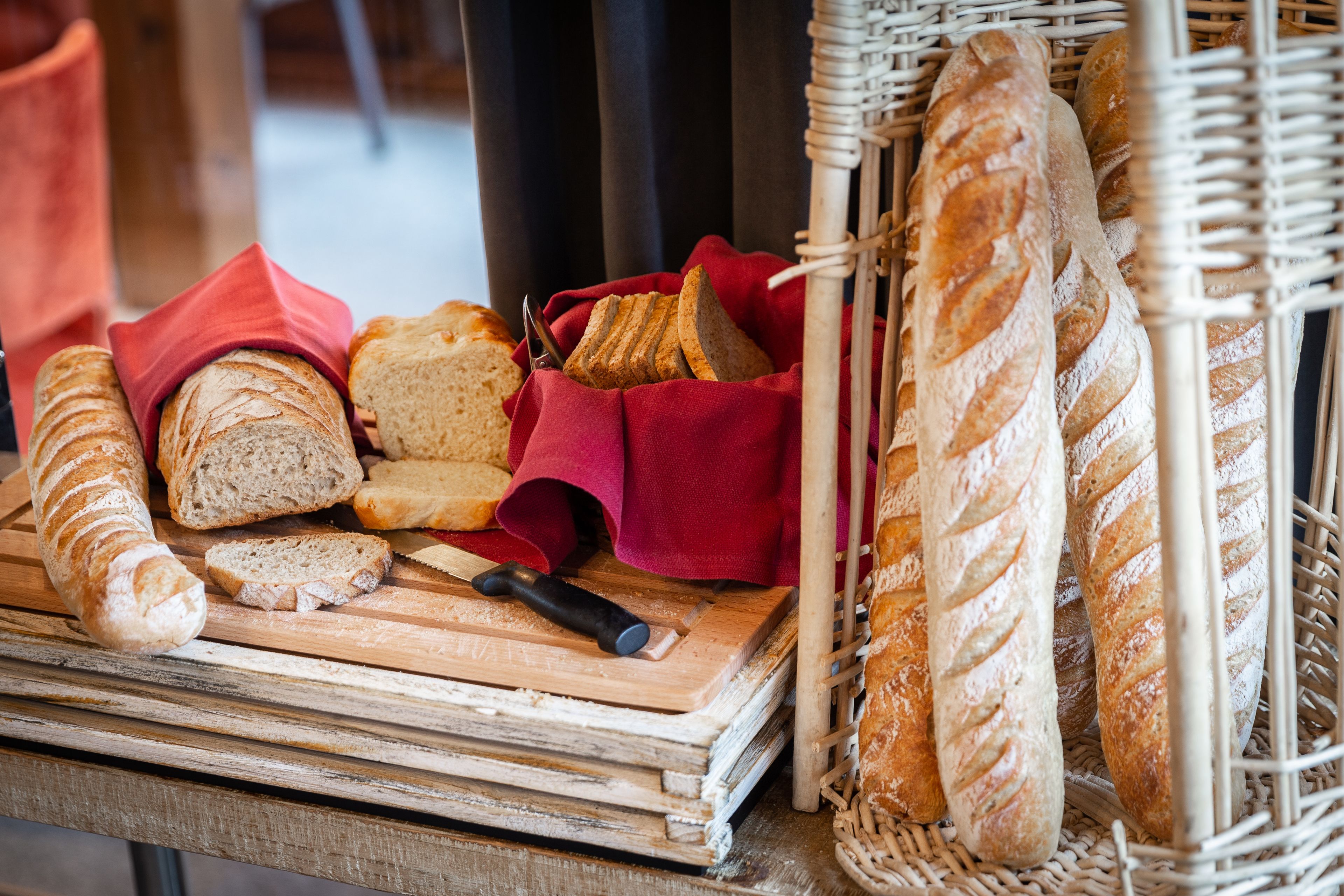 A variety of breads on a wooden cutting board, some in a wicker basket, and a red cloth nearby.