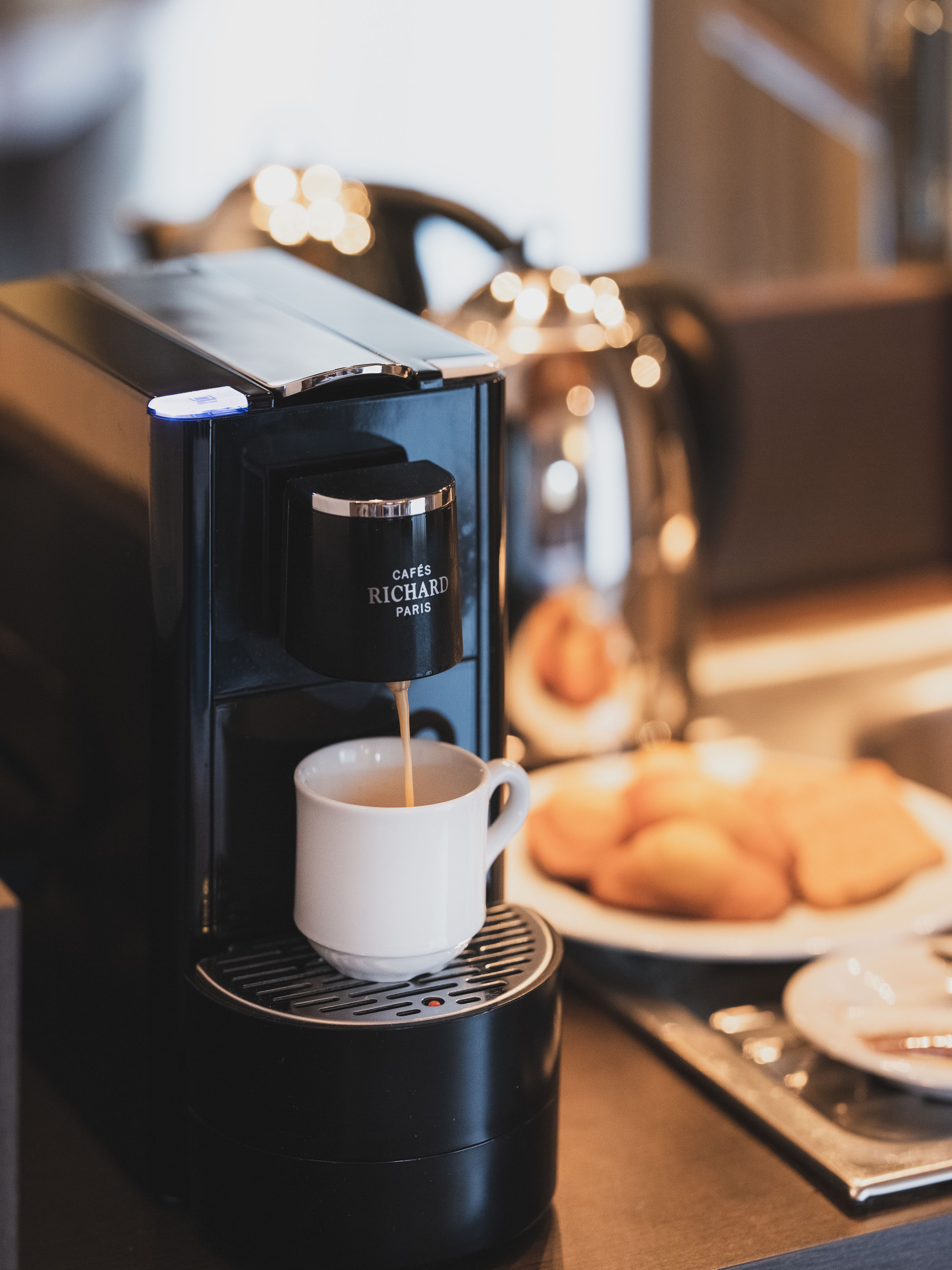 Una máquina de café sirviendo una taza, un plato de pasteles y un fondo desenfocado con luces.