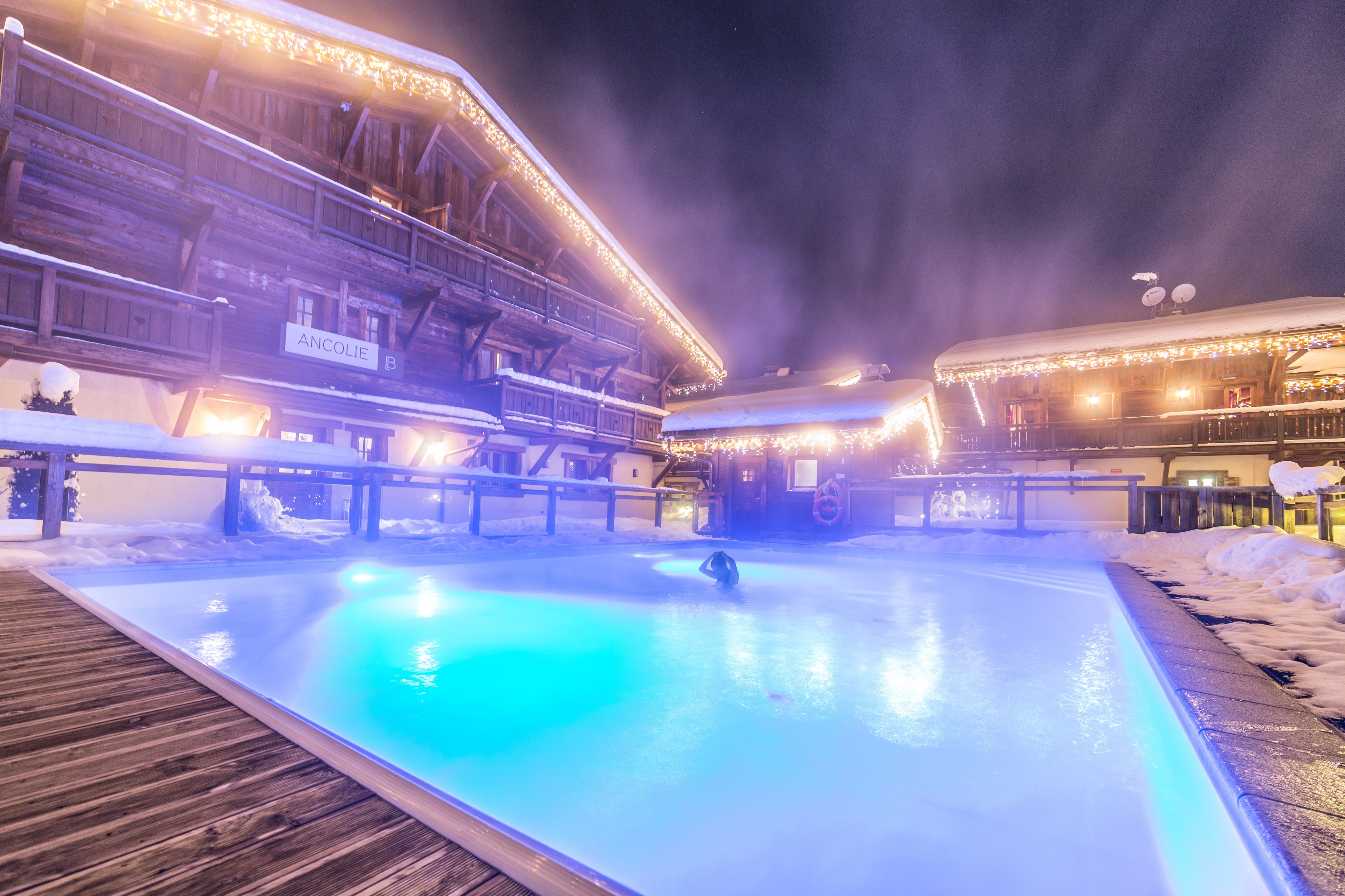 A heated outdoor pool with wooden deck, surrounded by snow-covered ground and buildings with illuminated railings.