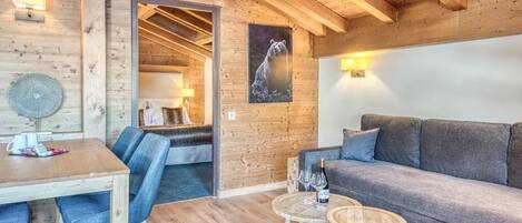 A cozy room with a wooden ceiling, a sofa, a dining table with chairs, and a small round table with a bottle and glasses.