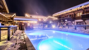 A snow-covered outdoor pool area with illuminated blue lights, wooden buildings with balconies, and snow on the ground. - Les Loges Blanches (Megeve)