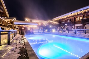 A snow-covered outdoor pool area with illuminated blue lights, wooden buildings with balconies, and snow on the ground. - Les Loges Blanches (Megeve)