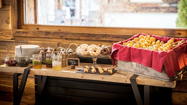 A breakfast buffet with pastries, jams, and honey-glazed bread, set against a wooden backdrop with a view of a building outside. - Les Loges Blanches (Megeve)