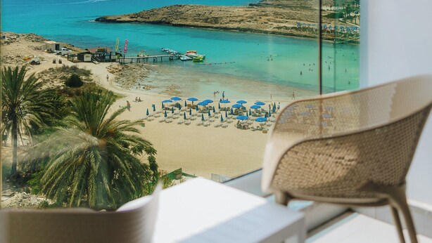 A balcony with a view of a beach, a pier, and a distant building. - Tasia Maris Sands - Adults Only (Ayia Napa)