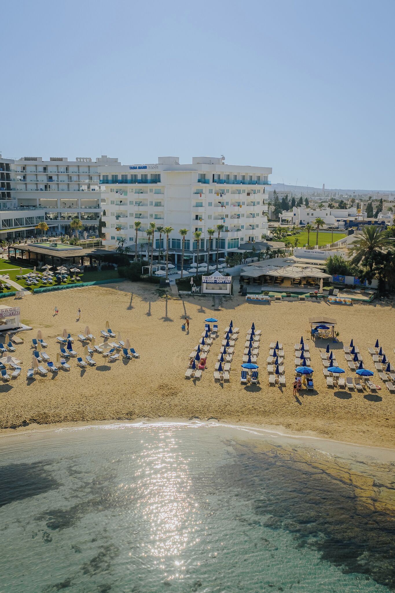 A beach resort with multiple levels of buildings, a sandy beach with rows of sun loungers, and a clear view of the ocean.