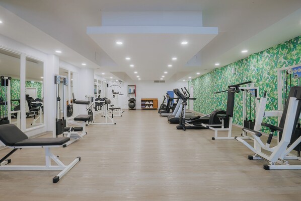Un gimnasio con diverso equipo de ejercicio, incluyendo cintas de correr y máquinas de pesas, dispuesto en un espacio amplio con suelo de madera y paredes de color verde con estampado.