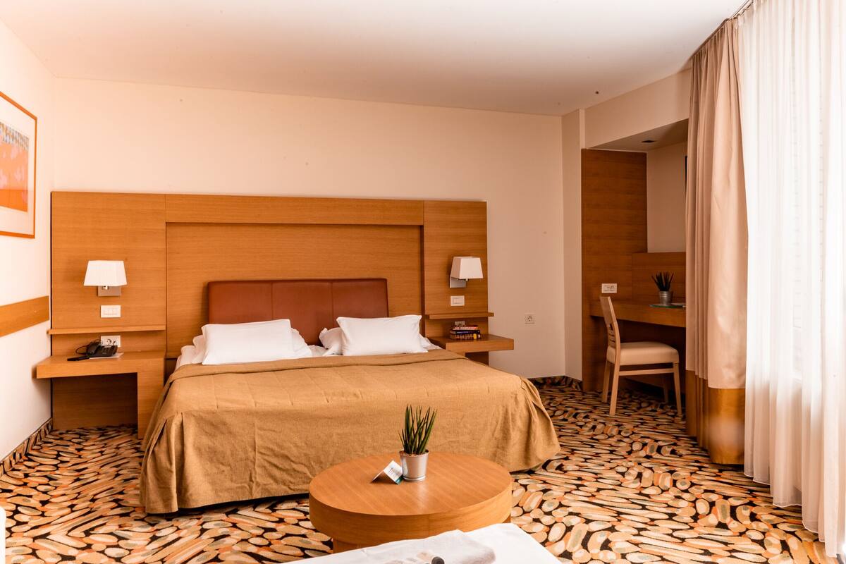 Ein Hotelzimmer mit einem großen Bett, zwei Nachttischen, einem runden Holztisch, einem Sessel und einem Schreibtisch mit Lampe.