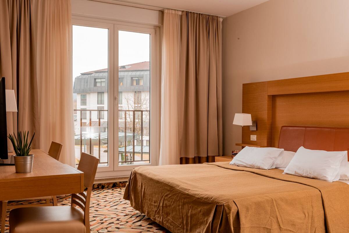 Ein Hotelzimmer mit einem Bett, einem Schreibtisch, einem Sessel, einer Zimmerpflanze, einem Fenster mit Vorhängen und Blick auf Gebäude.