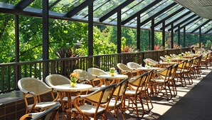 A covered outdoor seating area with wicker chairs and tables, surrounded by glass panels and greenery. - Hotel Šmarjeta - Terme Krka (Smarjeske Toplice)