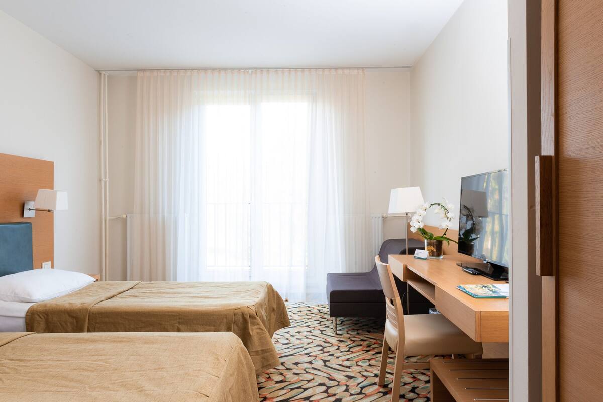 Ein Hotelzimmer mit zwei Betten, einem Schreibtisch, einem Stuhl, einem Fernseher und einem Fenster mit Vorhängen.
