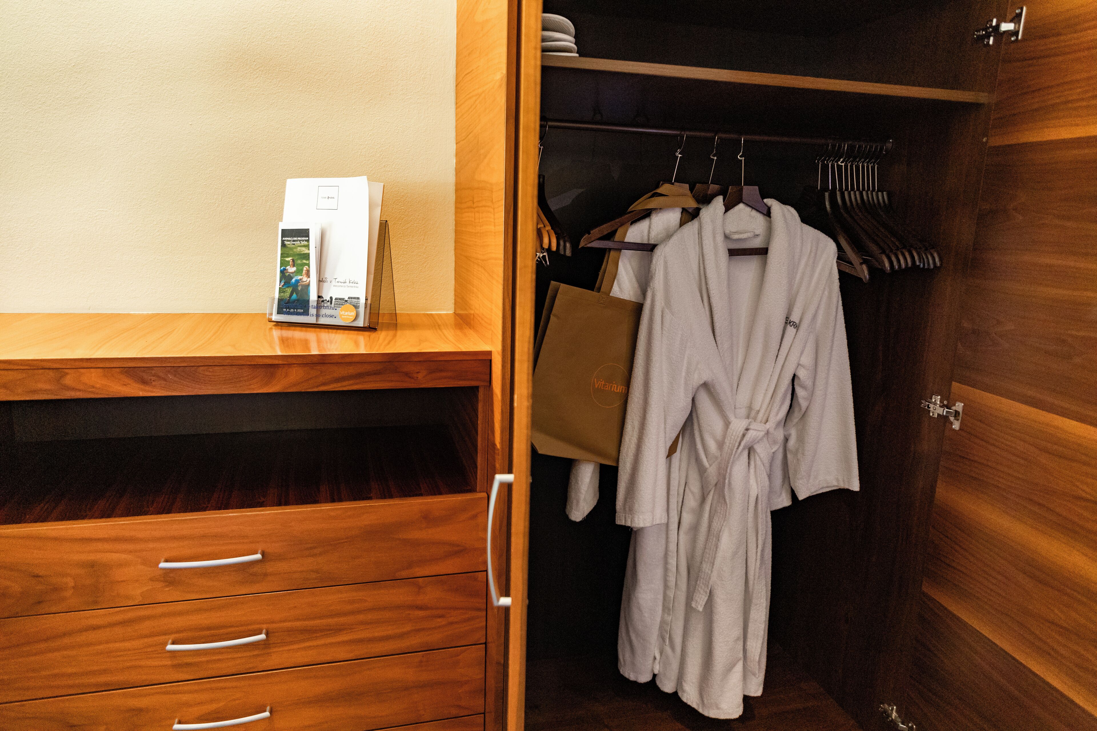 A wooden wardrobe with a white robe hanging on a rack, a brown bag, and a magazine on the shelf.
