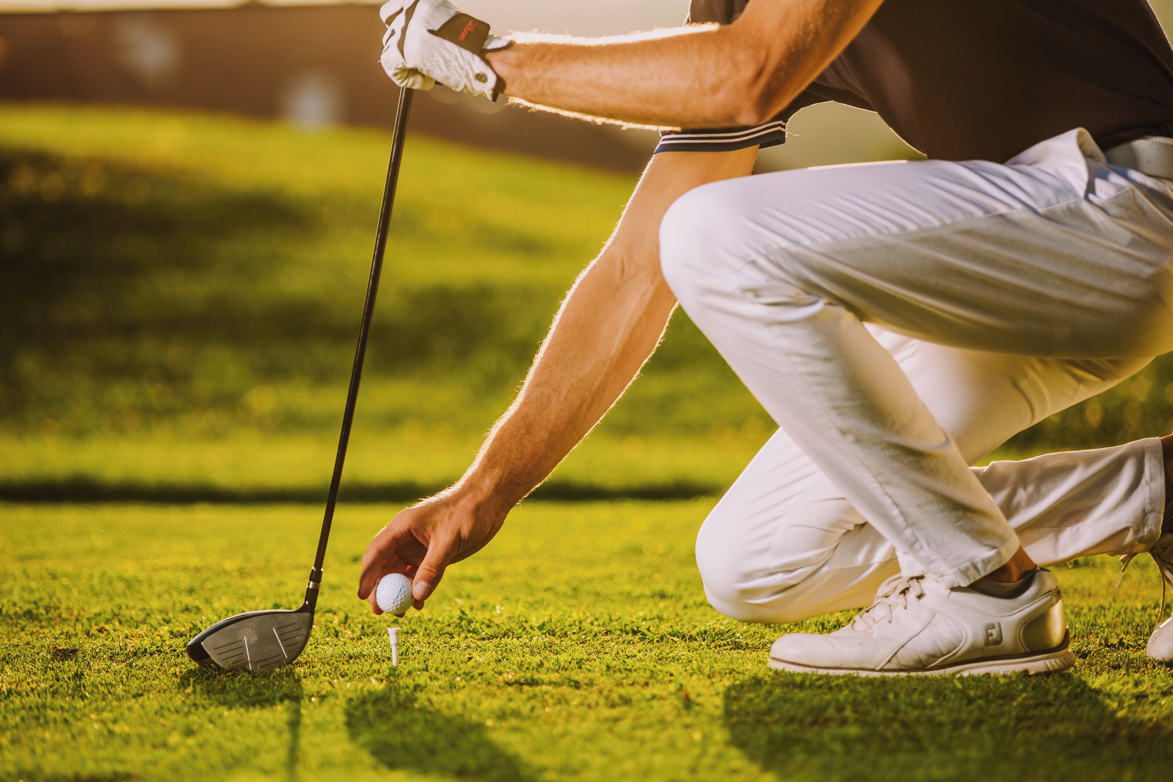 Une personne vêtue d’une chemise et d’un pantalon blancs, s’apprête à placer une balle de golf sur un support.