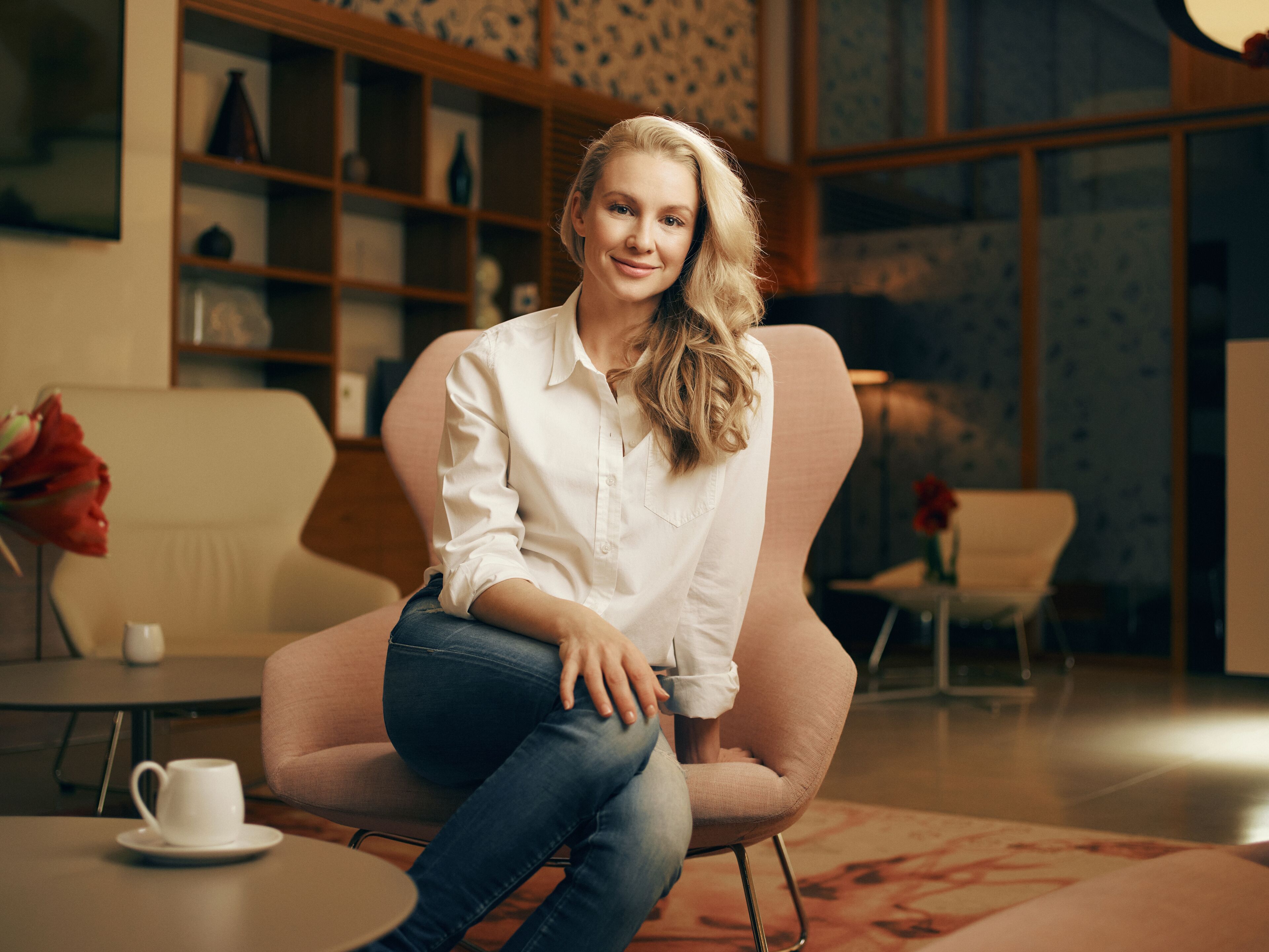 A woman seated on a chair in a room with a bookshelf, a table with a cup, and a vase with flowers.