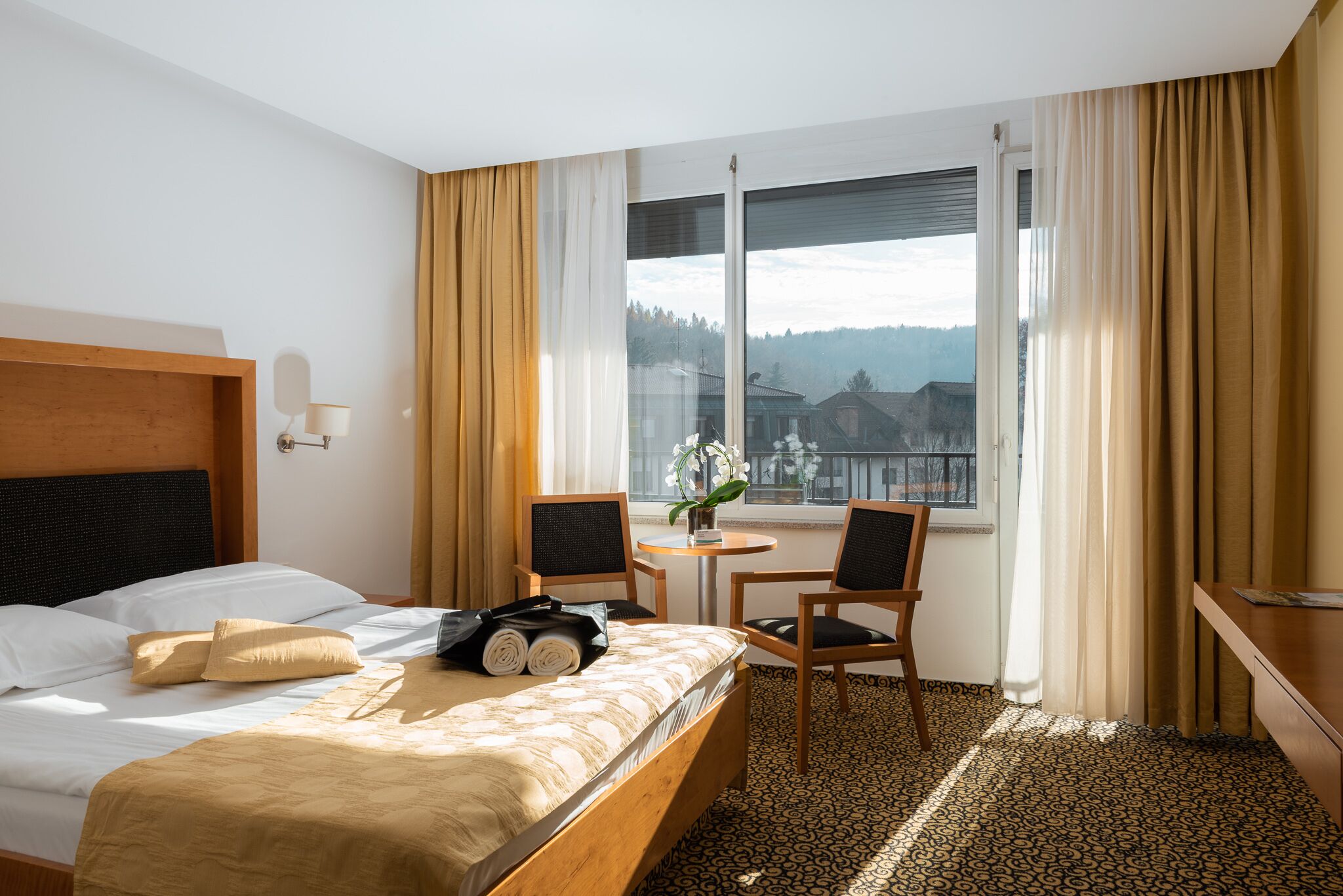 A hotel room with a bed, two chairs, a small table, and a window with curtains.