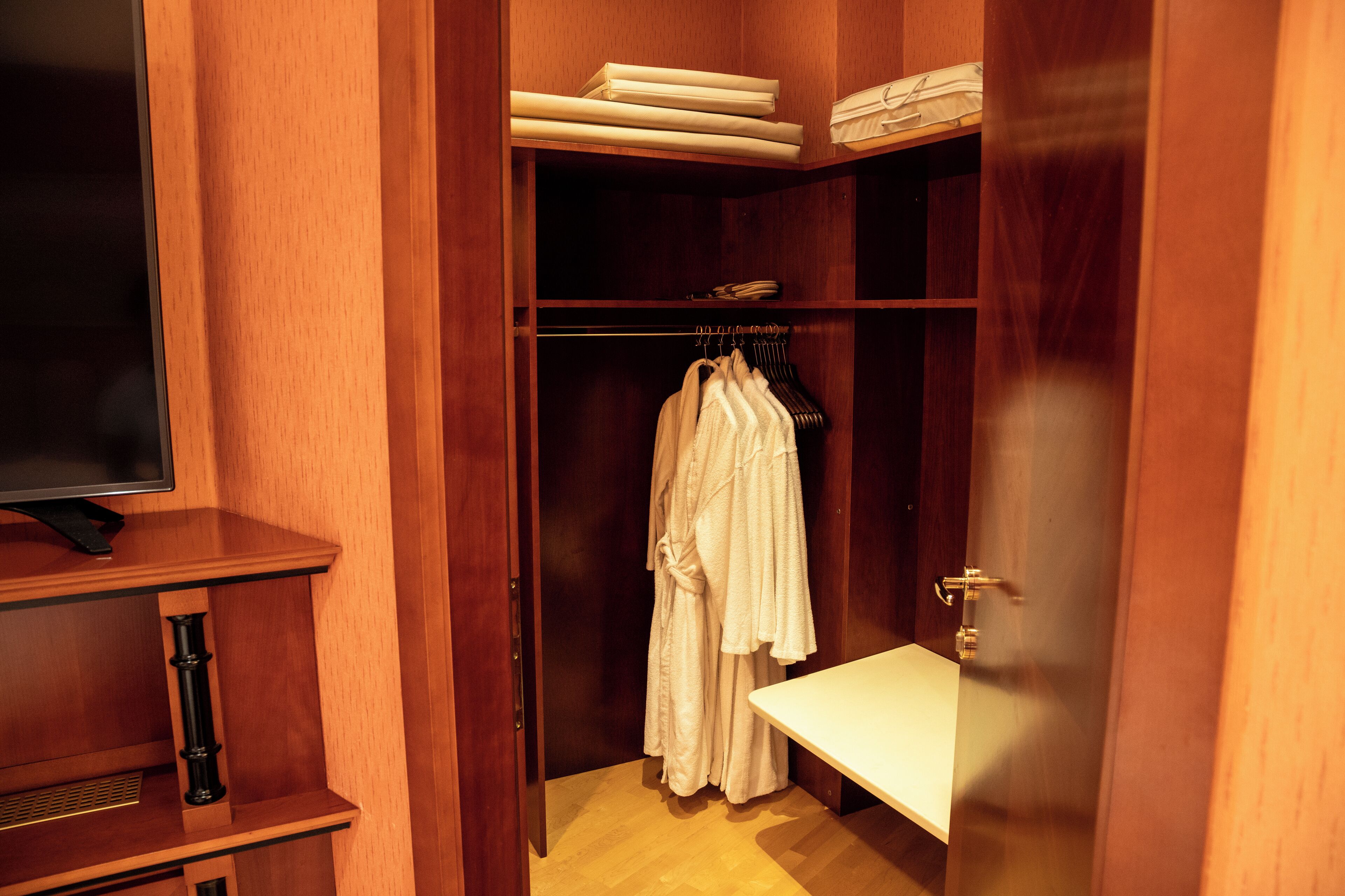 A wooden closet with hanging robes, a shelf with folded towels, and a bench.