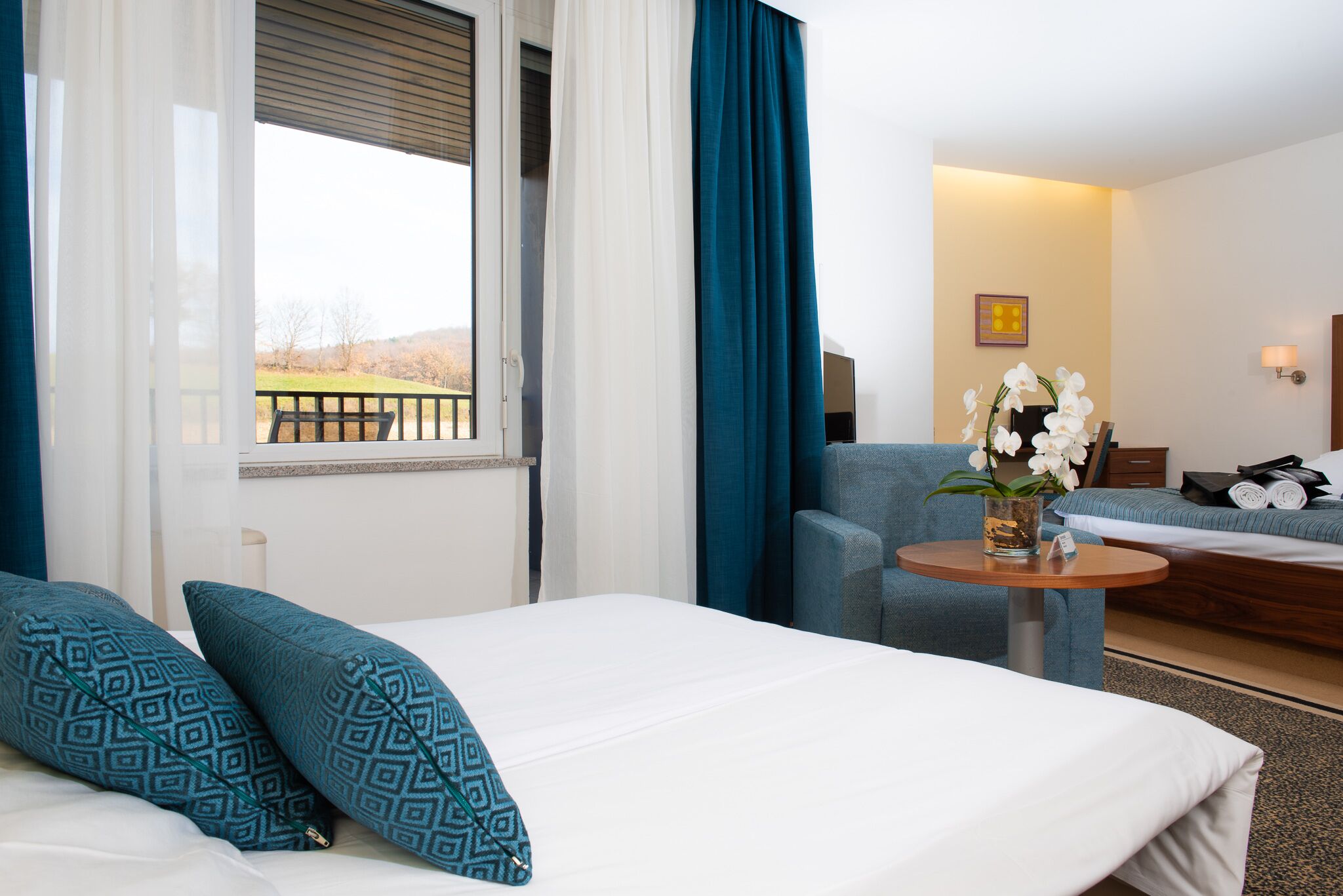 Habitación de hotel con cama, una mesita redonda con un florero, un sofá y vistas a un balcón con barandilla.