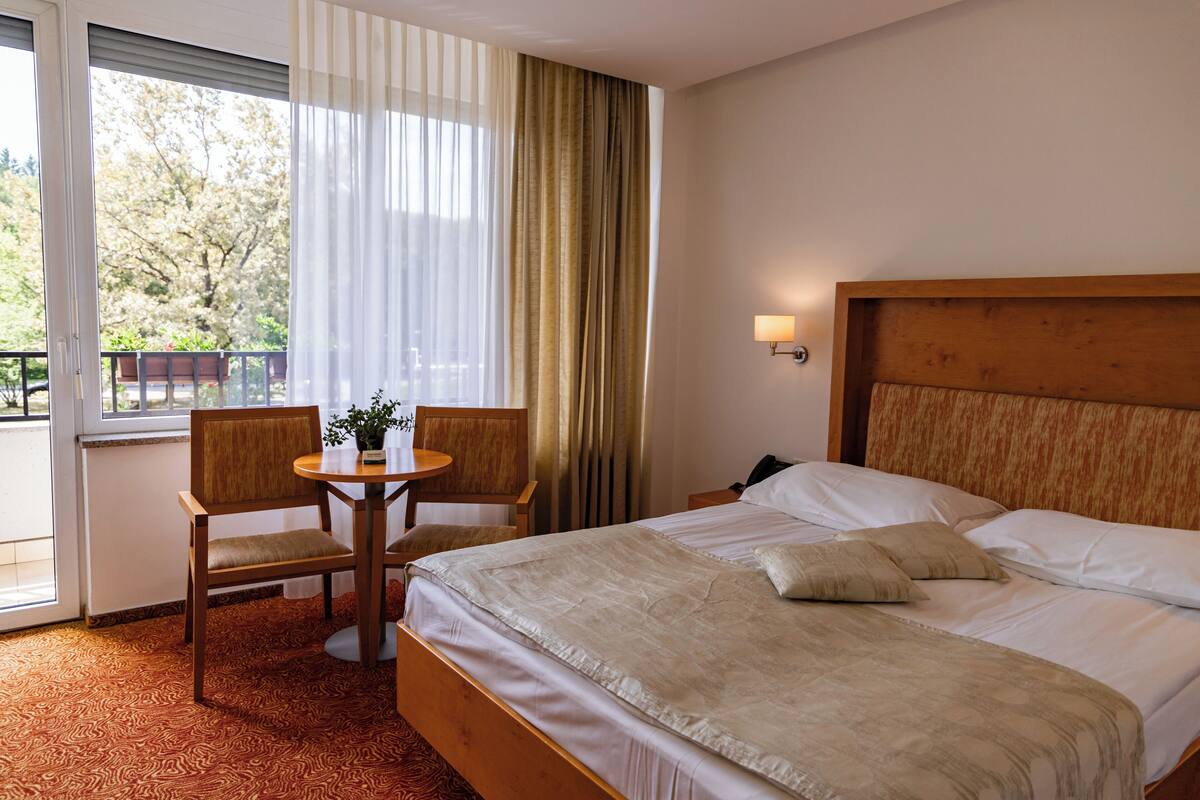 Une chambre d’hôtel avec un grand lit, deux fauteuils, une petite table avec une plante et une vue sur les arbres par la fenêtre.
