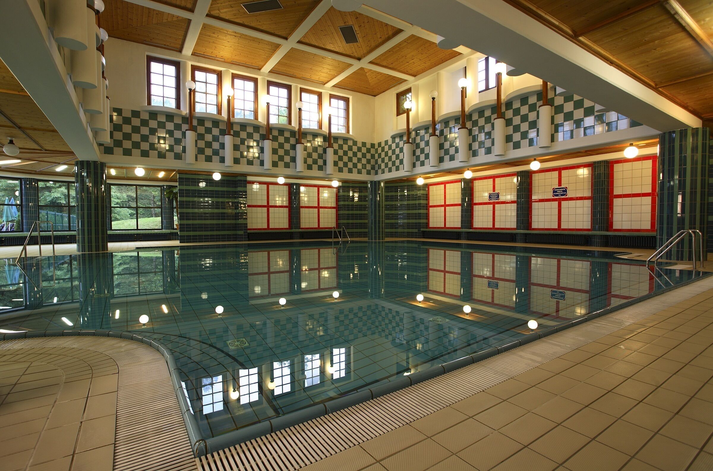 Une piscine intérieure aux murs à carreaux et au sol carrelé, entourée de fenêtres et surmontée d’un plafond en bois.