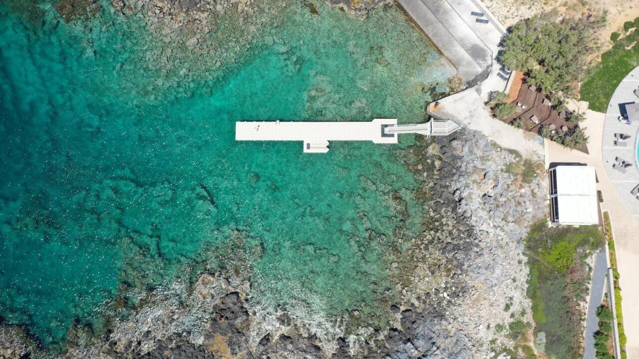 An aerial view of a coastal area with a white pier extending into turquoise waters, rocky terrain, and a small building with a paved area nearby.