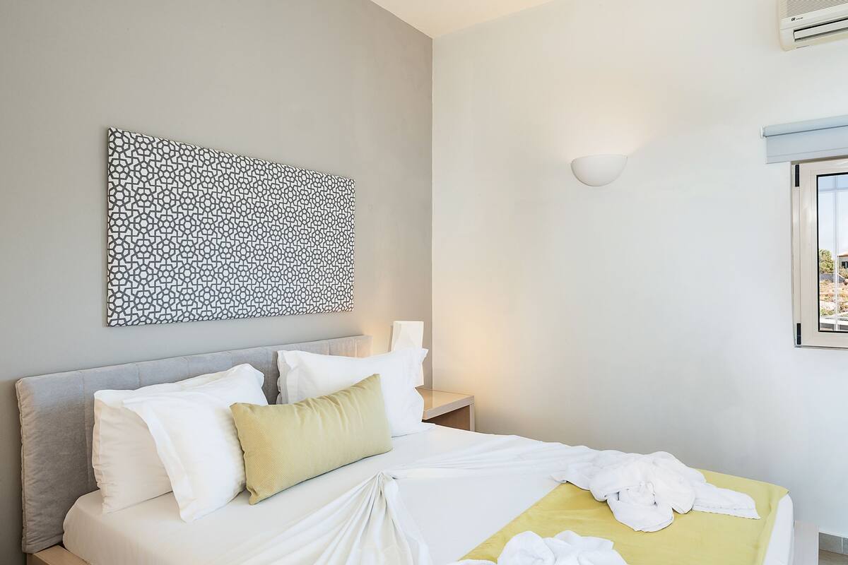 Una cama bien tendida con ropa de cama blanca y amarilla, un cabecero gris y una obra de arte abstracta colgada en la pared.
