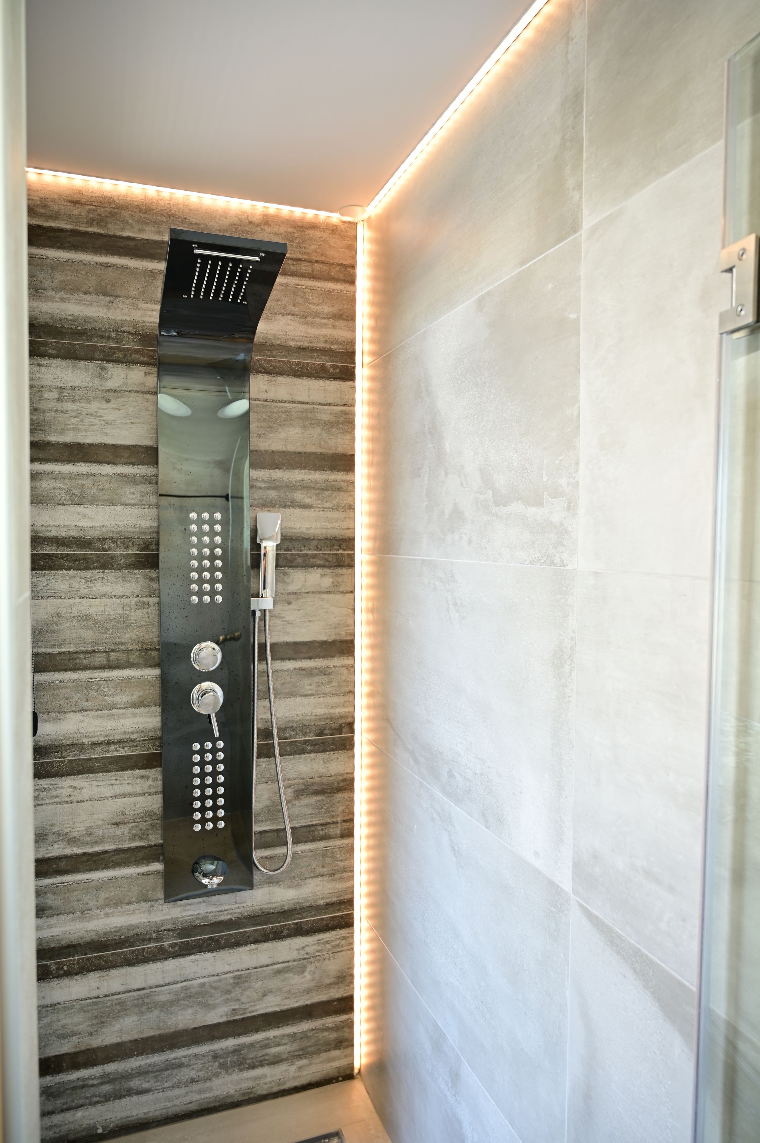 A modern shower with a control panel and a handheld showerhead, set against a textured wall with a warm light strip.