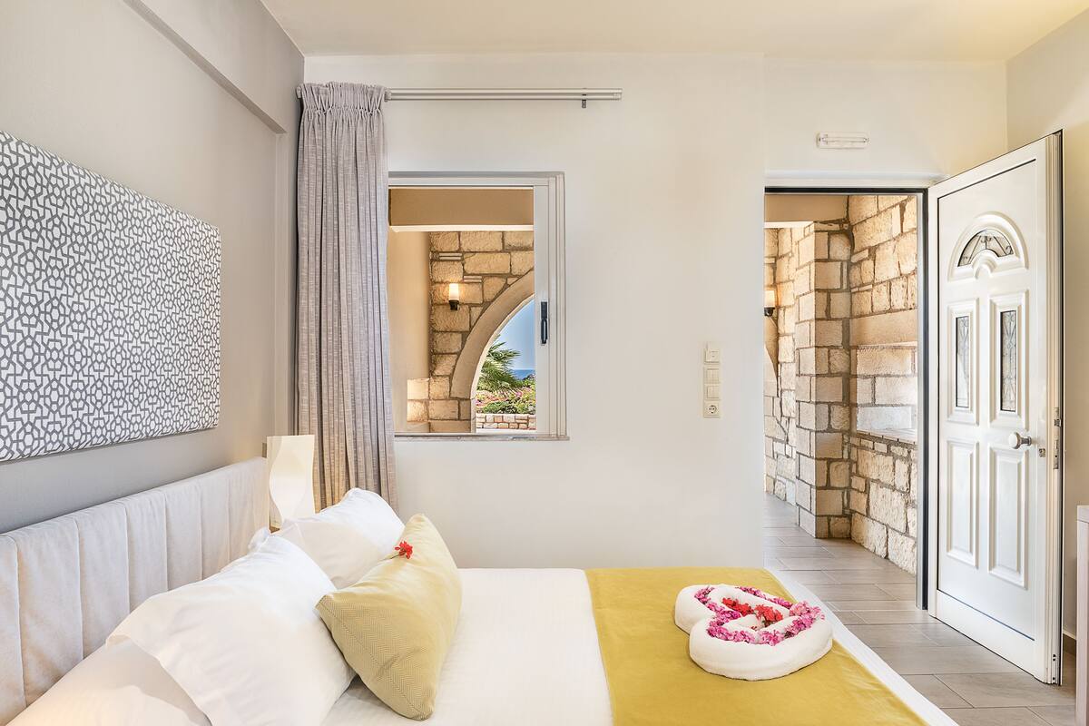 Habitación de hotel con cama, ventana con vista a un jardín, pared de piedra y una puerta.