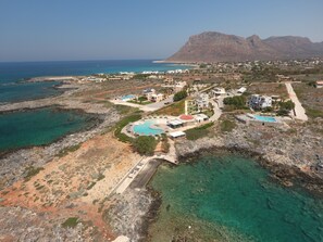 An aerial view of a coastal resort with a central swimming pool, multiple buildings, and a mountainous backdrop. - Kavos Hotel (Chania)