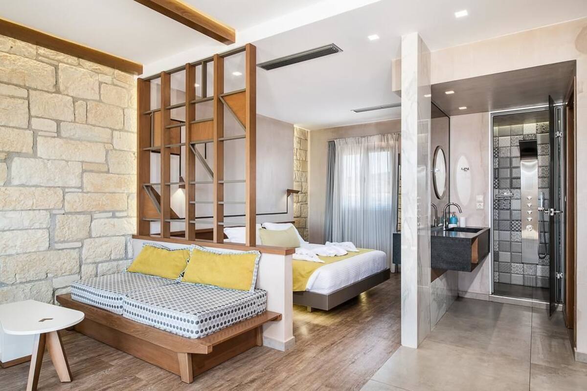 Habitación de hotel moderna con una pared de acento de piedra, un panel de madera, una cama con almohada amarilla, una mesita de noche y un baño con ducha y lavamanos.