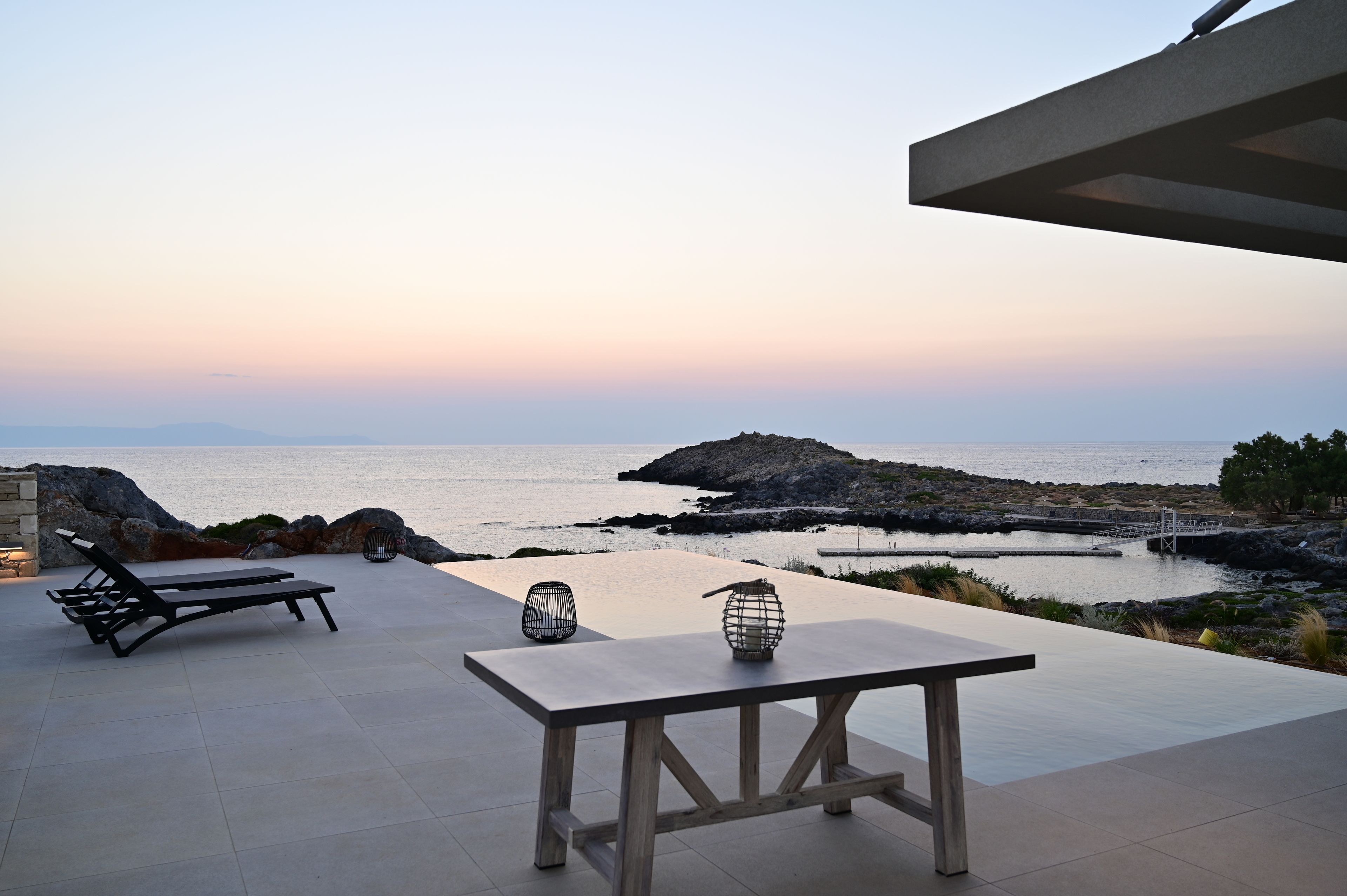 An outdoor patio with a table and two lounge chairs, overlooking a rocky coastline and calm sea.