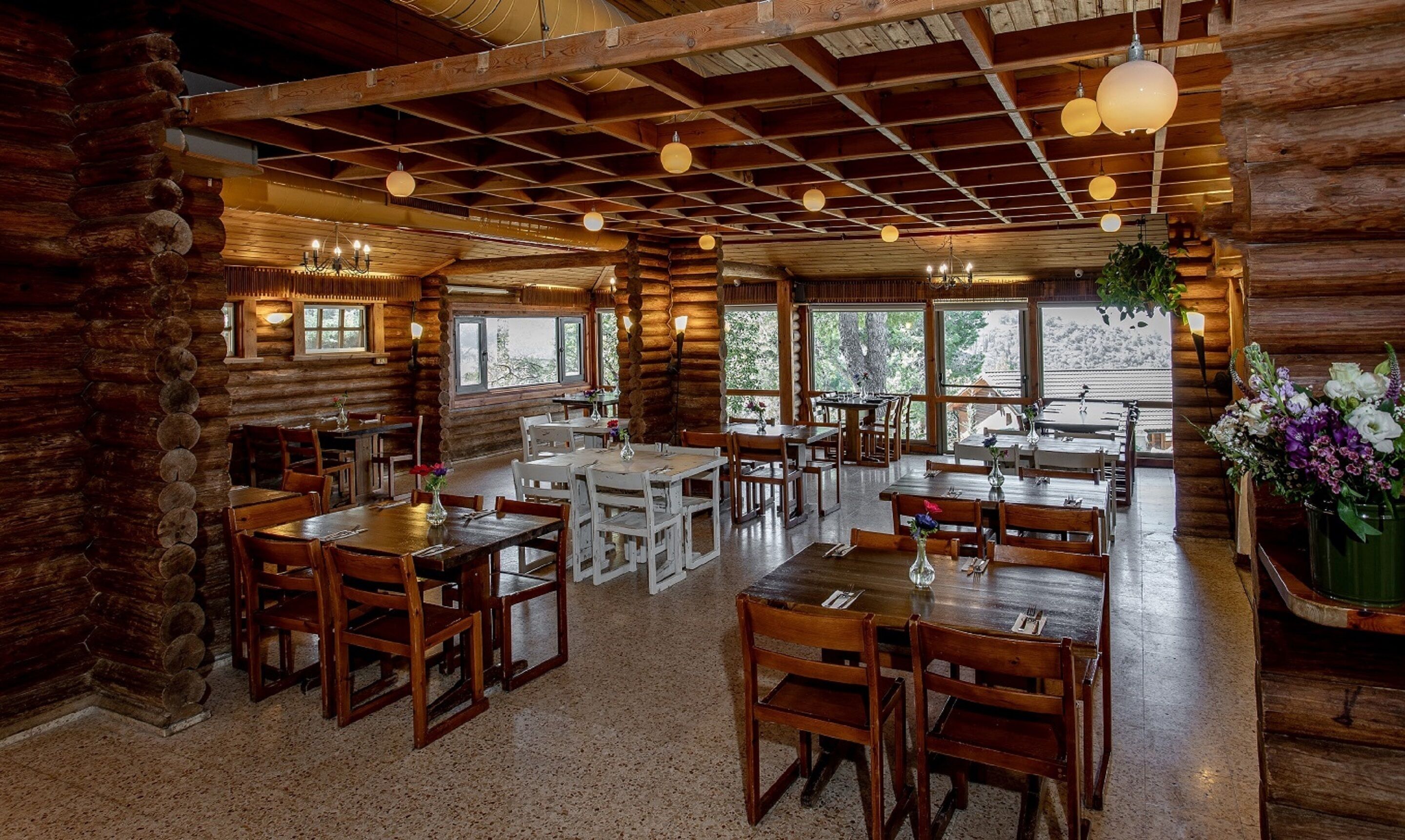 Interior rústico de una cabaña de troncos con múltiples mesas y sillas para comer, vigas de madera en el techo y amplias ventanas que ofrecen vistas al exterior.