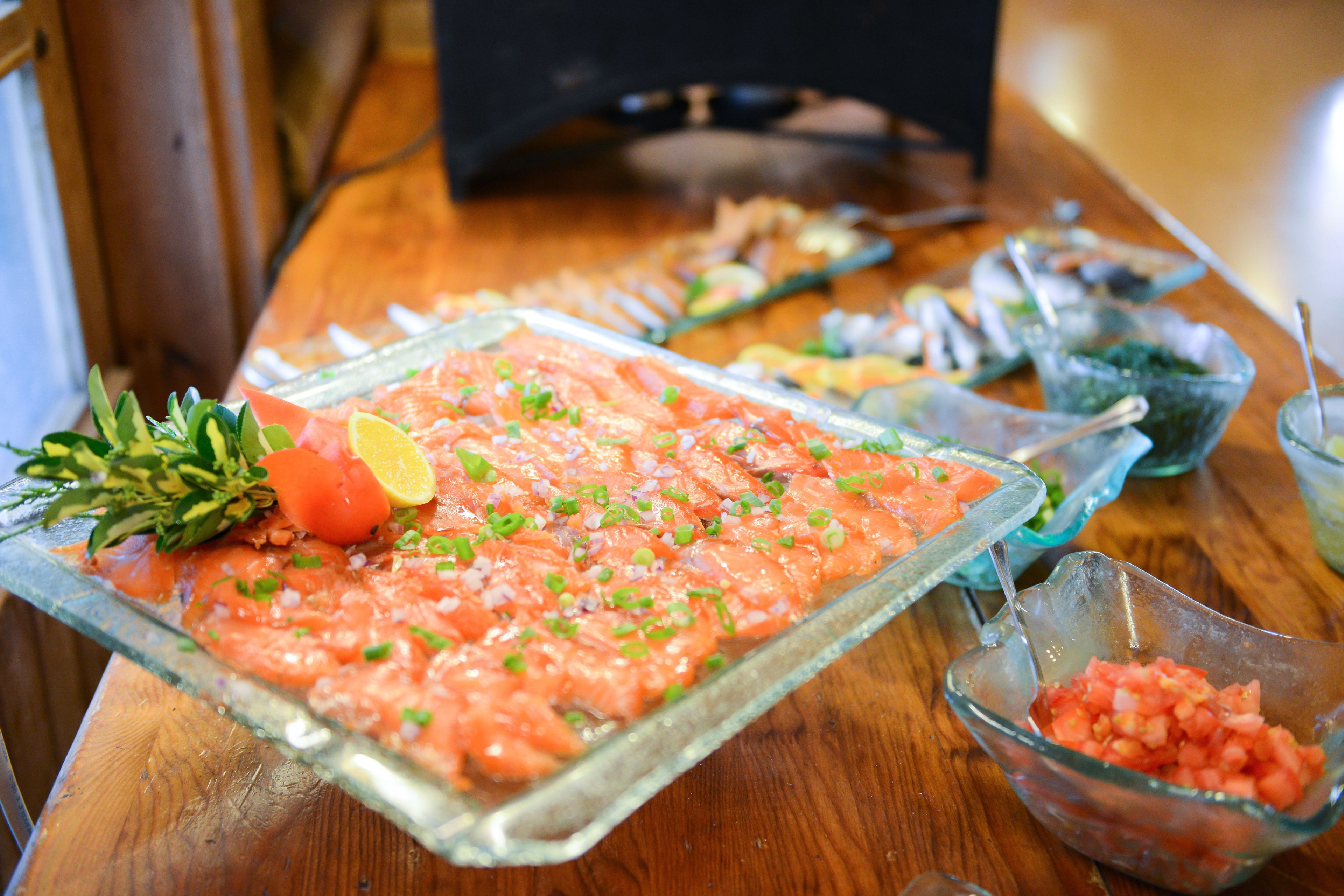 A glass dish with a raw fish salad garnished with herbs and lemon, surrounded by other food items on a wooden table.