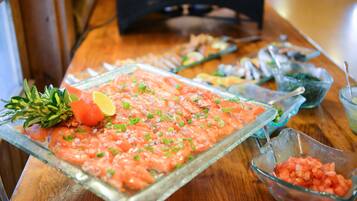 A glass dish with a raw fish salad garnished with herbs and lemon, surrounded by other food items on a wooden table.