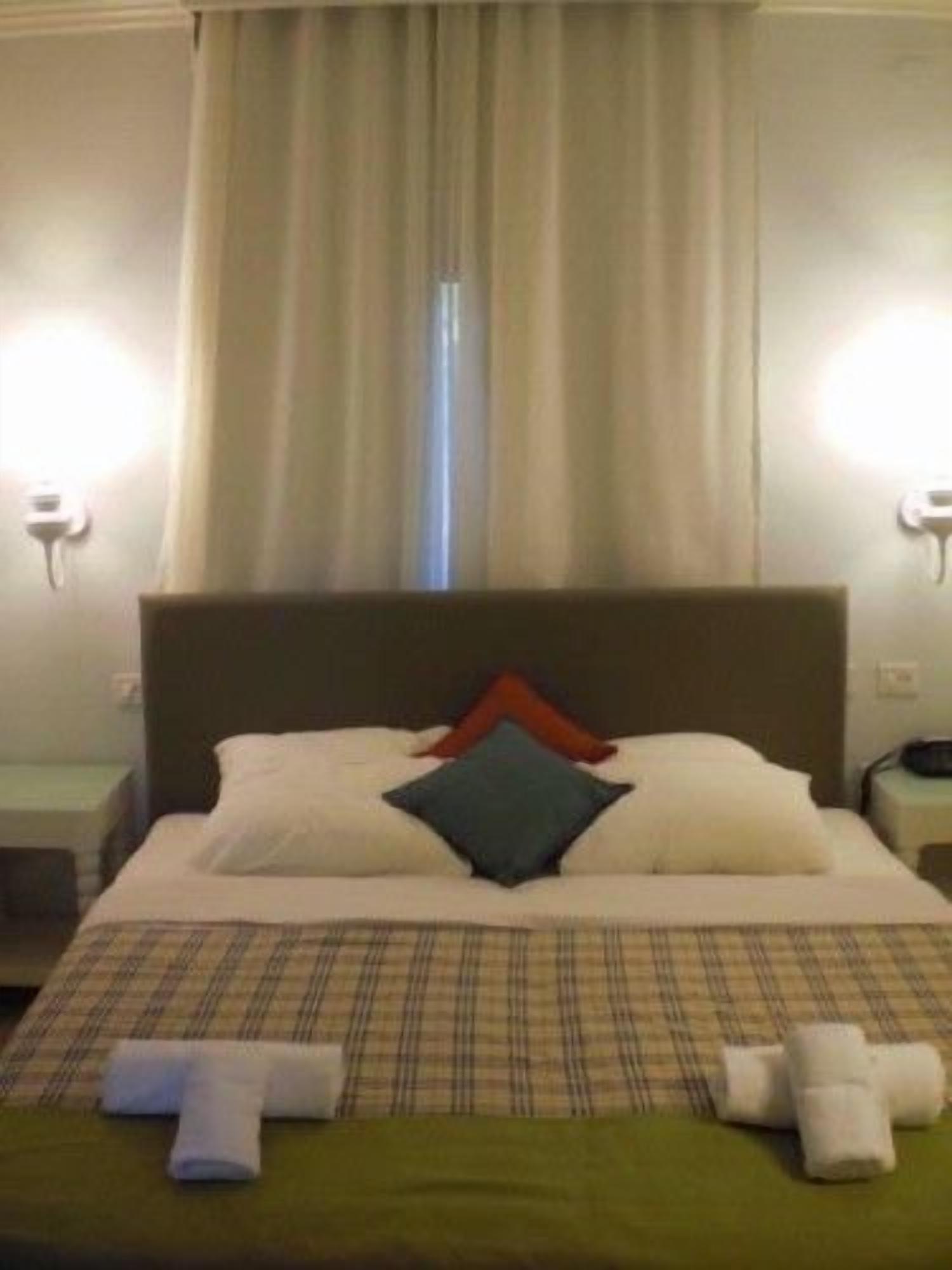 Una habitación de hotel con cama, mesita de noche, lámpara y ventana con cortinas.