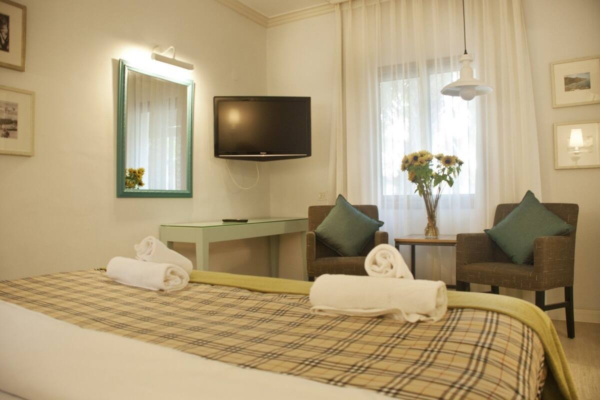Une chambre d’hôtel comprenant un lit, une télévision, deux fauteuils, une petite table avec un vase de fleurs et un miroir.