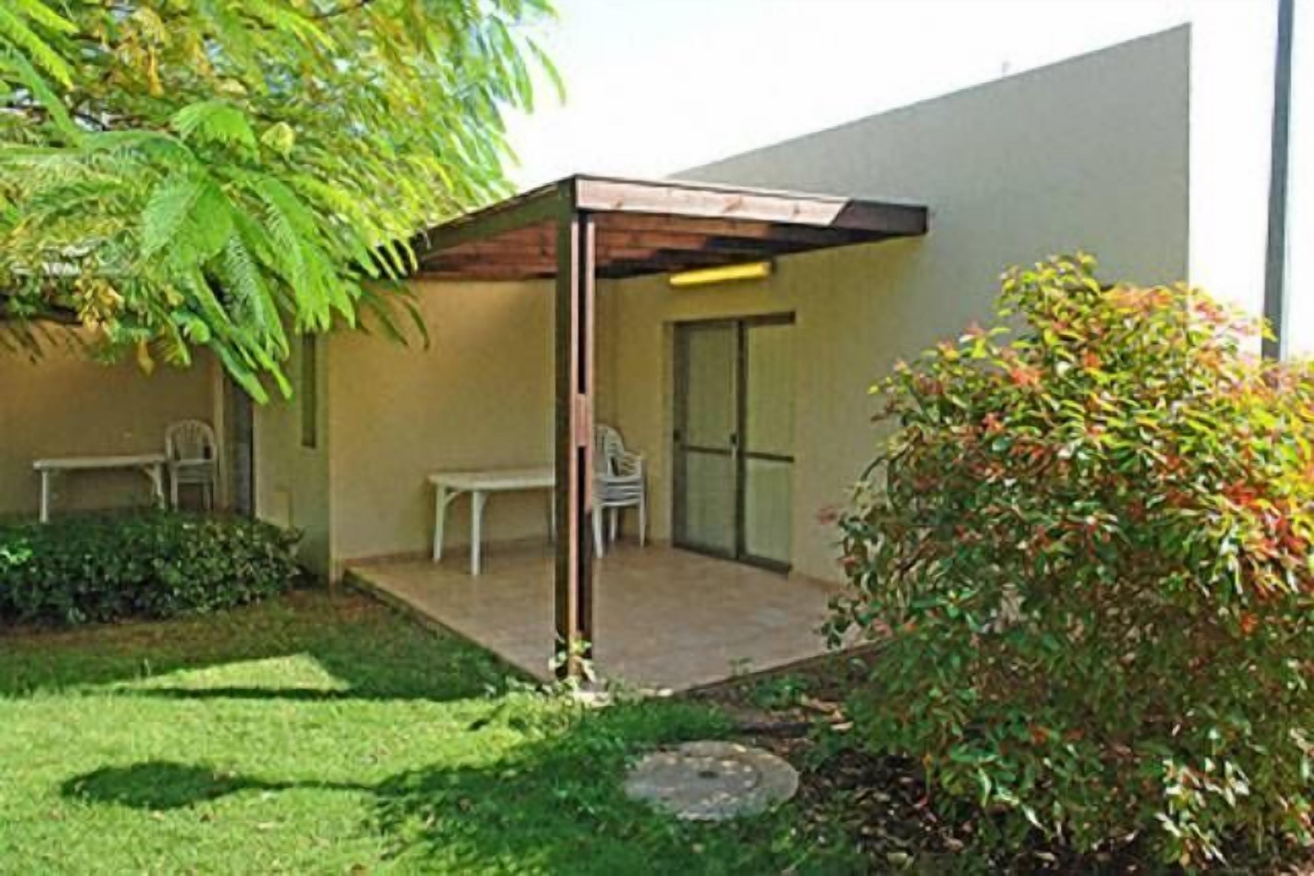 Un patio techado con una mesa y sillas, un pequeño jardín y un edificio con puerta de vidrio.