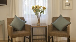Een woonkamer met twee gestreepten fauteuils, een kleine tafel met een vaas zonnebloemen, een aan de muur bevestigde televisie en een raam met transparante gordijnen.