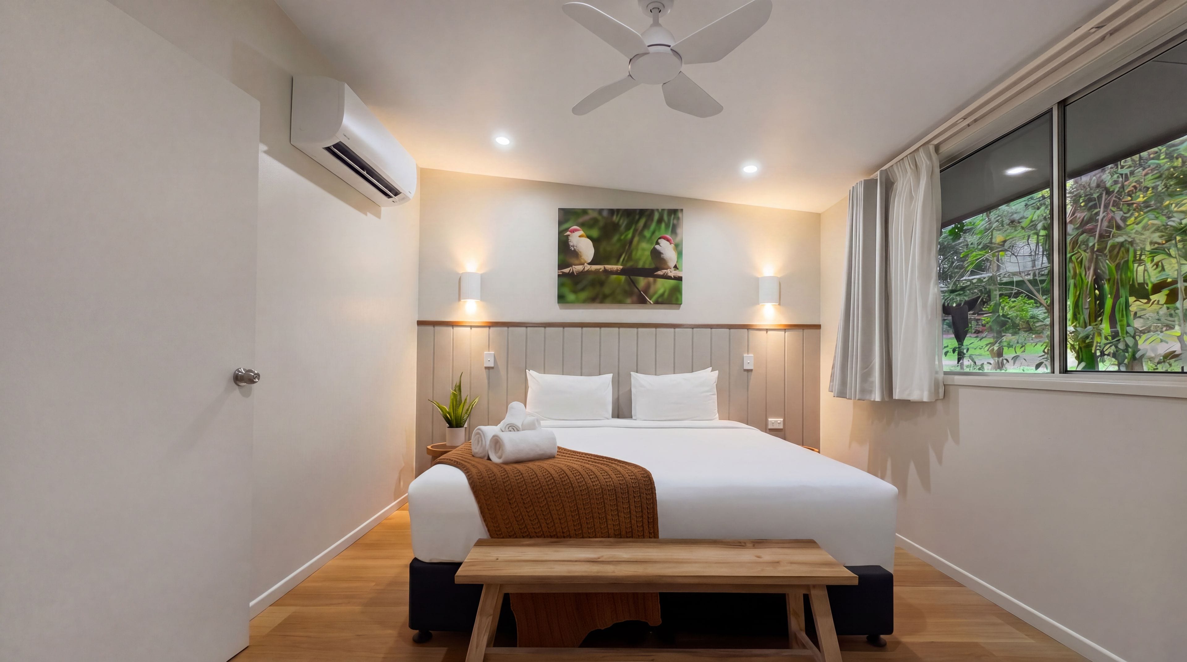 Et hotelværelse med seng, sengebord, aircondition, loftventilator, vægmonteret lampe, vindue med gardiner og et billede af fugle hængende på væggen.