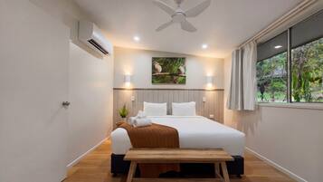Et hotelværelse med seng, sengebord, aircondition, loftventilator, vægmonteret lampe, vindue med gardiner og et billede af fugle hængende på væggen.