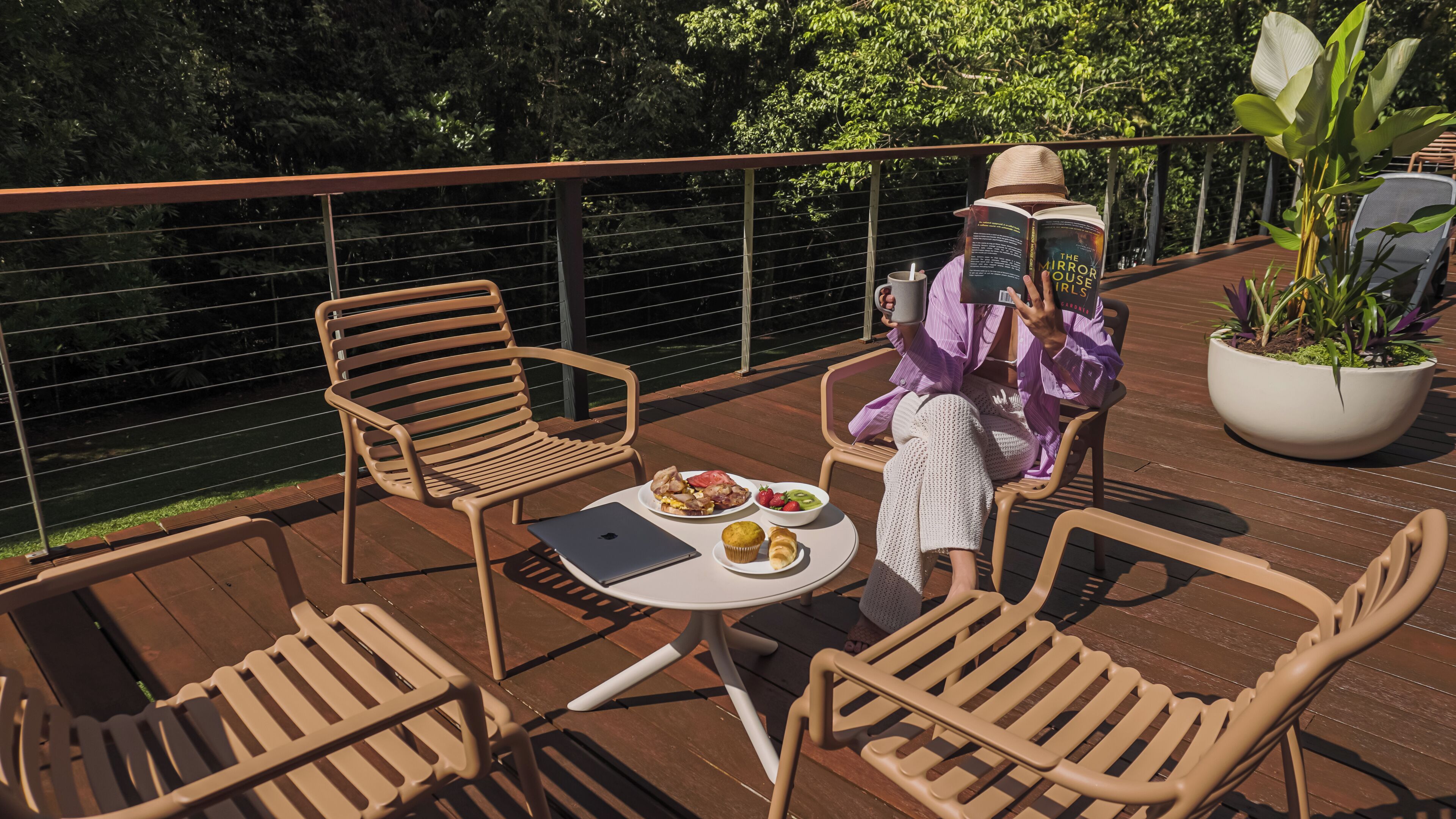 A person reading a book on a deck with a table set with food and a laptop.