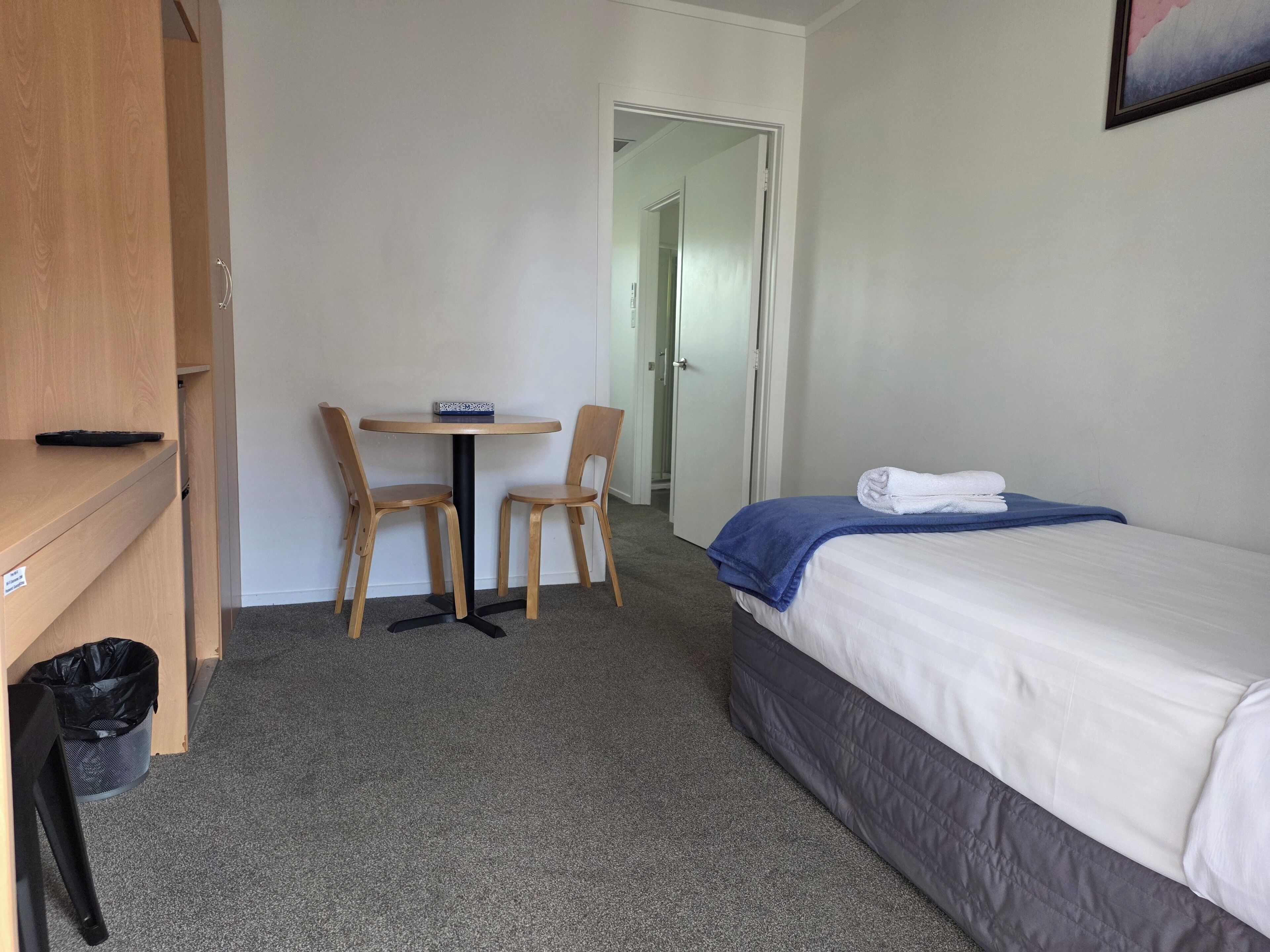 A hotel room with a bed, a small table with two chairs, a trash bin, and a door leading to another room.