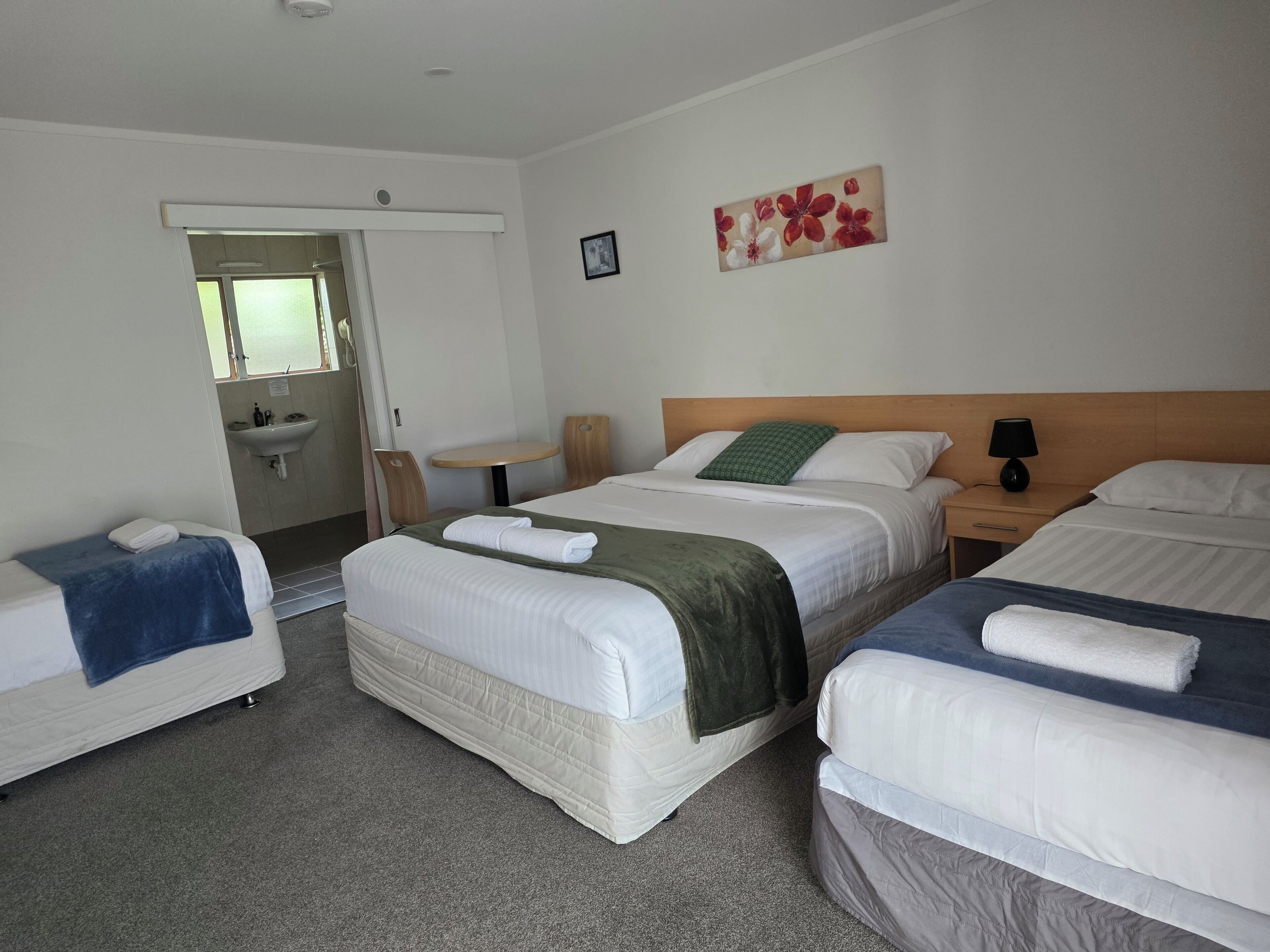 Habitación de hotel con tres camas, una mesita, una silla, un lavamanos y un cuadro en la pared.