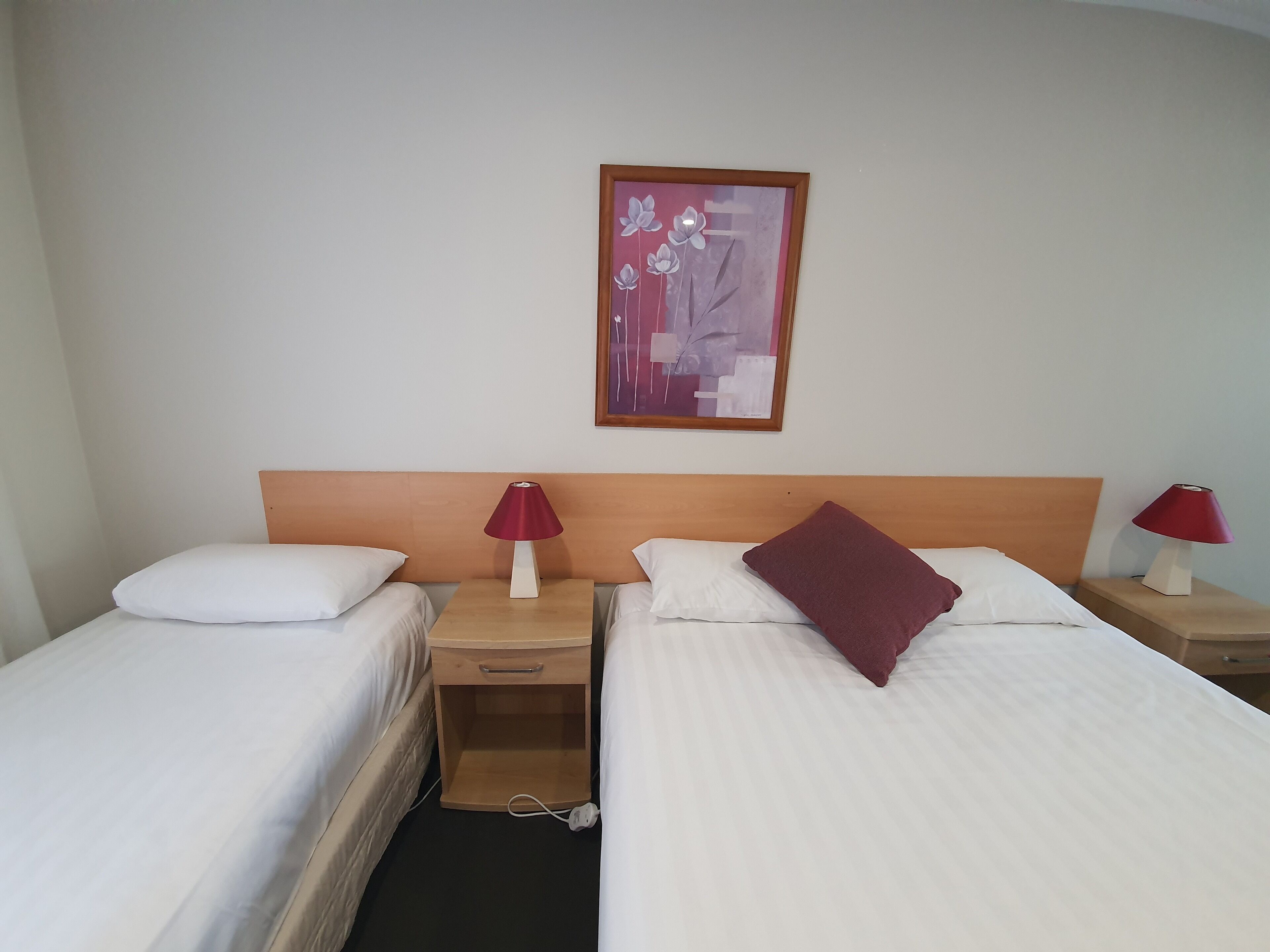 A hotel room with two single beds, each with white bedding and a red pillow. There is a wooden nightstand with a lamp between the beds, and a small wooden drawer. A framed artwork hangs on the wall above the beds.